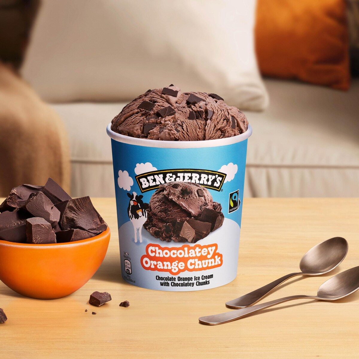 A tub and bowl of ice cream sitting on a couch with two spoons next to them